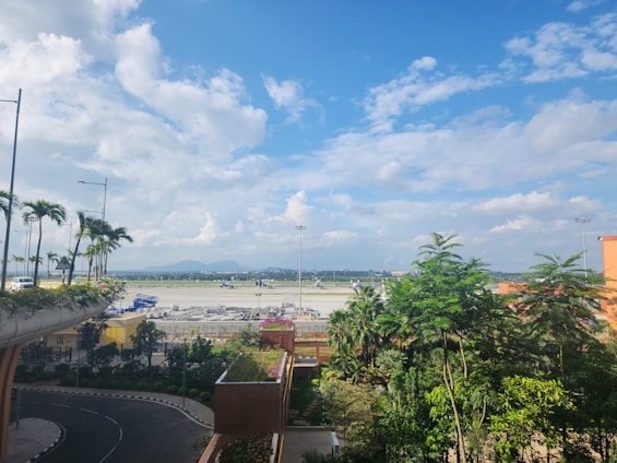 A corner NA plot with a paved road access and nearby greenery under a clear sunny sky.