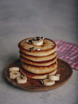 A stack of golden-brown pancakes is neatly arranged on a wooden plate. Slices of bananas and small pieces of chocolate are artfully scattered on top of the pancakes and around the plate, creating a pleasing presentation. A pink and white checkered napkin is partially visible in the background, adding a touch of color.