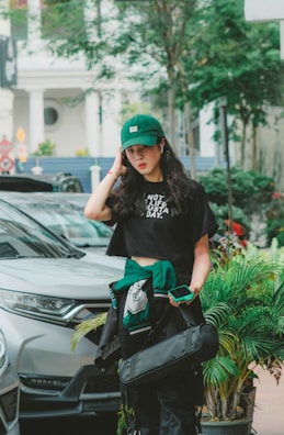 A young person with long hair stands outdoors, wearing a green cap and a black t-shirt that reads 'Not a lifestyle, a mustaplay'. They have a casual appearance, with a phone in hand and a jacket around their waist. A silver vehicle, green plants, and urban structures are visible in the background.