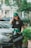 A young person with long hair stands outdoors, wearing a green cap and a black t-shirt that reads 'Not a lifestyle, a mustaplay'. They have a casual appearance, with a phone in hand and a jacket around their waist. A silver vehicle, green plants, and urban structures are visible in the background.