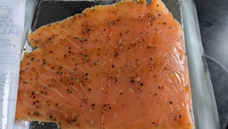 A large piece of raw, seasoned salmon fillet sits on a transparent surface. The fish has a vibrant orange color with visible striations and is sprinkled with black pepper and various seasonings. To the side, there is a partially visible product packaging with text.