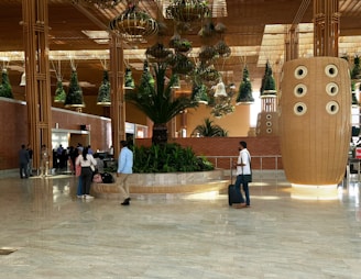 A spacious indoor area with a modern architectural design, featuring large hanging plant installations and brick walls. There are several people walking or standing around, some carrying luggage. A prominent, large decorative structure with circular elements stands to the right side.