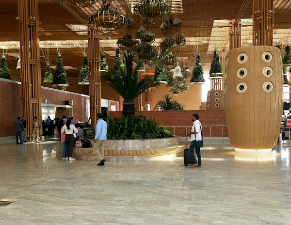 A spacious indoor area with a modern architectural design, featuring large hanging plant installations and brick walls. There are several people walking or standing around, some carrying luggage. A prominent, large decorative structure with circular elements stands to the right side.