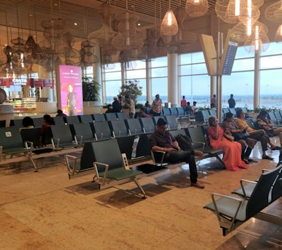 The scene is set in a modern airport terminal with several rows of empty and occupied black metal seating. There are people sitting, and some walking around. Toward the back, there's a Costa Coffee shop, and to the right, a large digital advertisement screen. The terminal has large ceiling lights and wicker pendant lights hanging. The expansive windows at the back reveal a view of the airport tarmac and the outside landscape.