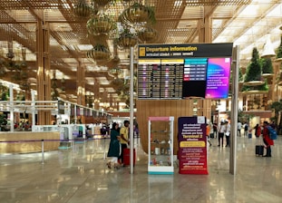 A spacious airport terminal with a high ceiling featuring hanging greenery and wooden structures. The central focus is a large departure information board displaying flight details. Several people are visible, some checking information, others walking with luggage. The terminal emits a modern and organized atmosphere.