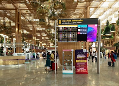 A spacious airport terminal with a high ceiling featuring hanging greenery and wooden structures. The central focus is a large departure information board displaying flight details. Several people are visible, some checking information, others walking with luggage. The terminal emits a modern and organized atmosphere.