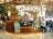 A tea shop named Chaayos located in an airport. The kiosk features a modern and inviting design with wood paneling and greenery. A variety of teas and snacks are displayed in glass cases. People are seen interacting with the shop, with travel bags in tow. The environment is bustling and well-lit, typical of an airport setting.