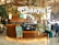 A tea shop named Chaayos located in an airport. The kiosk features a modern and inviting design with wood paneling and greenery. A variety of teas and snacks are displayed in glass cases. People are seen interacting with the shop, with travel bags in tow. The environment is bustling and well-lit, typical of an airport setting.