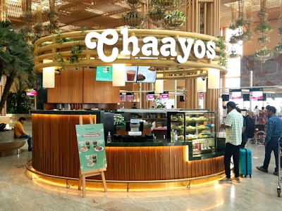 A tea shop named Chaayos located in an airport. The kiosk features a modern and inviting design with wood paneling and greenery. A variety of teas and snacks are displayed in glass cases. People are seen interacting with the shop, with travel bags in tow. The environment is bustling and well-lit, typical of an airport setting.