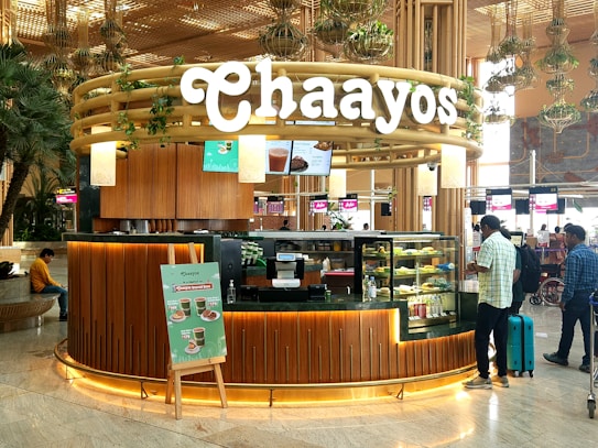 A tea shop named Chaayos located in an airport. The kiosk features a modern and inviting design with wood paneling and greenery. A variety of teas and snacks are displayed in glass cases. People are seen interacting with the shop, with travel bags in tow. The environment is bustling and well-lit, typical of an airport setting.