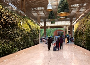 A spacious indoor area with lush vertical gardens covering the walls. Hanging plant installations are suspended from the ceiling, and several people are walking with suitcases on a polished marble floor.