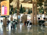 A busy airport terminal with modern architectural design features wooden columns and green plants hanging from the ceiling. There is a prominent digital advertisement display for Amazon with a woman in traditional attire. People are walking with luggage, creating an active and bustling atmosphere.