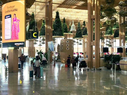 A busy airport terminal with modern architectural design features wooden columns and green plants hanging from the ceiling. There is a prominent digital advertisement display for Amazon with a woman in traditional attire. People are walking with luggage, creating an active and bustling atmosphere.