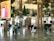 A busy airport terminal with modern architectural design features wooden columns and green plants hanging from the ceiling. There is a prominent digital advertisement display for Amazon with a woman in traditional attire. People are walking with luggage, creating an active and bustling atmosphere.