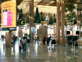 A busy airport terminal with modern architectural design features wooden columns and green plants hanging from the ceiling. There is a prominent digital advertisement display for Amazon with a woman in traditional attire. People are walking with luggage, creating an active and bustling atmosphere.