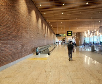 A spacious airport terminal corridor with a brick wall on the left, a moving walkway in the center, and a right-side seating area with large windows. The ceiling features wooden panels with spotlights, and overhead signs indicate gates D8 and D9. Several passengers, including one with a backpack, walk through the terminal.