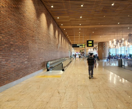 A spacious airport terminal corridor with a brick wall on the left, a moving walkway in the center, and a right-side seating area with large windows. The ceiling features wooden panels with spotlights, and overhead signs indicate gates D8 and D9. Several passengers, including one with a backpack, walk through the terminal.