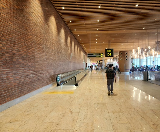 A spacious airport terminal corridor with a brick wall on the left, a moving walkway in the center, and a right-side seating area with large windows. The ceiling features wooden panels with spotlights, and overhead signs indicate gates D8 and D9. Several passengers, including one with a backpack, walk through the terminal.