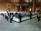 A spacious airport security check area with a long counter. Several people stand in line, some waiting while others interact with security staff. The environment features high ceilings, large glass windows, and wooden architectural elements. The floor is tiled, reflecting the natural light coming from the windows.