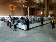 A spacious airport security check area with a long counter. Several people stand in line, some waiting while others interact with security staff. The environment features high ceilings, large glass windows, and wooden architectural elements. The floor is tiled, reflecting the natural light coming from the windows.