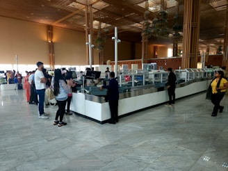 A spacious airport security check area with a long counter. Several people stand in line, some waiting while others interact with security staff. The environment features high ceilings, large glass windows, and wooden architectural elements. The floor is tiled, reflecting the natural light coming from the windows.