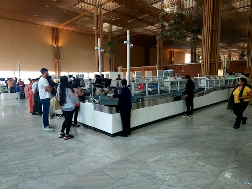 A spacious airport security check area with a long counter. Several people stand in line, some waiting while others interact with security staff. The environment features high ceilings, large glass windows, and wooden architectural elements. The floor is tiled, reflecting the natural light coming from the windows.