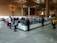 A spacious airport security check area with a long counter. Several people stand in line, some waiting while others interact with security staff. The environment features high ceilings, large glass windows, and wooden architectural elements. The floor is tiled, reflecting the natural light coming from the windows.