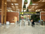 A modern airport terminal with an 'International departures' sign displayed prominently in multiple languages. The architectural design features a high ceiling with decorative plants hanging from above and polished stone flooring. Security personnel and travelers are visible near the entrance gates, and there is a blend of natural and artificial lighting.