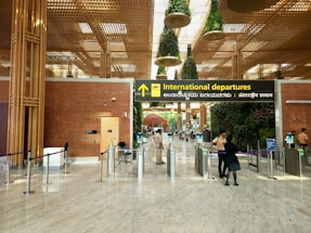 A modern airport terminal with an 'International departures' sign displayed prominently in multiple languages. The architectural design features a high ceiling with decorative plants hanging from above and polished stone flooring. Security personnel and travelers are visible near the entrance gates, and there is a blend of natural and artificial lighting.