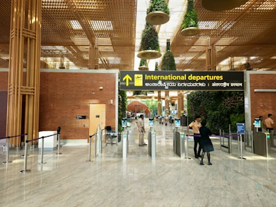A modern airport terminal with an 'International departures' sign displayed prominently in multiple languages. The architectural design features a high ceiling with decorative plants hanging from above and polished stone flooring. Security personnel and travelers are visible near the entrance gates, and there is a blend of natural and artificial lighting.