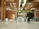A modern airport terminal with an 'International departures' sign displayed prominently in multiple languages. The architectural design features a high ceiling with decorative plants hanging from above and polished stone flooring. Security personnel and travelers are visible near the entrance gates, and there is a blend of natural and artificial lighting.
