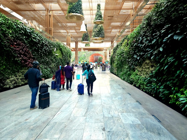 A group of people with suitcases walk through a spacious corridor lined with lush vertical gardens on both sides. The ceiling features hanging greenery and wooden lattice structures, creating an inviting and modern atmosphere.