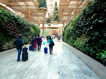 A group of people with suitcases walk through a spacious corridor lined with lush vertical gardens on both sides. The ceiling features hanging greenery and wooden lattice structures, creating an inviting and modern atmosphere.