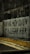 A graffiti on a concrete wall with a Spanish phrase written in large, bold black letters. The wall is part of an urban setting with a gritty texture, and the ground in front of it has sparse patches of grass and a sidewalk.