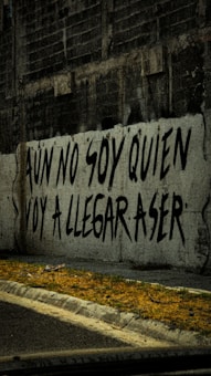 A graffiti on a concrete wall with a Spanish phrase written in large, bold black letters. The wall is part of an urban setting with a gritty texture, and the ground in front of it has sparse patches of grass and a sidewalk.