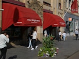 A storefront with red awnings displaying the name 'P.G.C. Hajenius, Tobacconist since 1826' is situated on a busy street. Several people are walking by, some holding shopping bags. In front of the store, there is a large planter with vibrant flowers and greenery. The building has intricate architectural features, including decorative stonework.