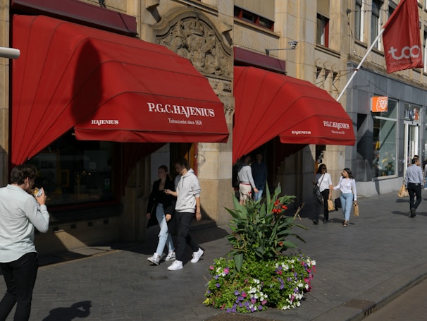 A storefront with red awnings displaying the name 'P.G.C. Hajenius, Tobacconist since 1826' is situated on a busy street. Several people are walking by, some holding shopping bags. In front of the store, there is a large planter with vibrant flowers and greenery. The building has intricate architectural features, including decorative stonework.