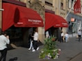 A storefront with red awnings displaying the name 'P.G.C. Hajenius, Tobacconist since 1826' is situated on a busy street. Several people are walking by, some holding shopping bags. In front of the store, there is a large planter with vibrant flowers and greenery. The building has intricate architectural features, including decorative stonework.