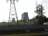 Several large cooling towers from a power plant are visible in the background beyond a green field, partially obscured by a tall electrical transmission tower in the foreground. Some trees and various other electrical infrastructure are also present.