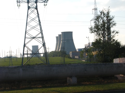 Several large cooling towers from a power plant are visible in the background beyond a green field, partially obscured by a tall electrical transmission tower in the foreground. Some trees and various other electrical infrastructure are also present.