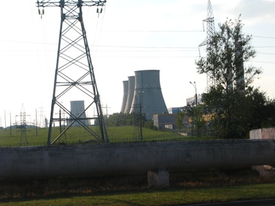Several large cooling towers from a power plant are visible in the background beyond a green field, partially obscured by a tall electrical transmission tower in the foreground. Some trees and various other electrical infrastructure are also present.