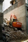 An orange excavator is performing demolition work amidst a pile of rubble and debris, surrounded by partially demolished buildings. The machine is branded with 'Hitachi' on its side and is set against a backdrop of brick and concrete structures.