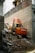 An orange excavator is performing demolition work amidst a pile of rubble and debris, surrounded by partially demolished buildings. The machine is branded with 'Hitachi' on its side and is set against a backdrop of brick and concrete structures.