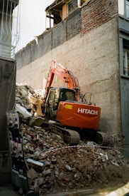 An orange excavator is performing demolition work amidst a pile of rubble and debris, surrounded by partially demolished buildings. The machine is branded with 'Hitachi' on its side and is set against a backdrop of brick and concrete structures.