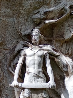A detailed stone carving of a warrior figure holding a sword with both hands, featuring intricate details such as a crown or helmet on the figure's head and a robe or cloak draped over its shoulders. The background includes additional carvings of natural elements and mythical creatures.