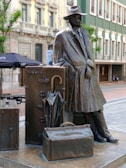 Bronze statue of a man wearing a hat and coat leaning against an arrangement of large suitcases and a folded umbrella. The backdrop includes an outdoor setting with cafe umbrellas, historical buildings, and a modern theater entrance labeled 'Filarmonica'.