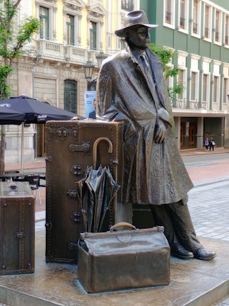 Bronze statue of a man wearing a hat and coat leaning against an arrangement of large suitcases and a folded umbrella. The backdrop includes an outdoor setting with cafe umbrellas, historical buildings, and a modern theater entrance labeled 'Filarmonica'.