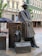 Bronze statue of a man wearing a hat and coat leaning against an arrangement of large suitcases and a folded umbrella. The backdrop includes an outdoor setting with cafe umbrellas, historical buildings, and a modern theater entrance labeled 'Filarmonica'.