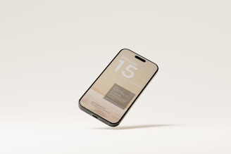 A smartphone is depicted floating against a plain, light-colored background. The screen displays text and graphics, suggesting a promotional or mock-up design for an iPhone 15.