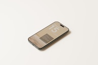 A smartphone lies on a light, neutral-colored surface. The screen displays text indicating 'iPhone 15' with a minimalistic design. The phone is oriented at a slight angle, allowing visibility of the top and side edges.